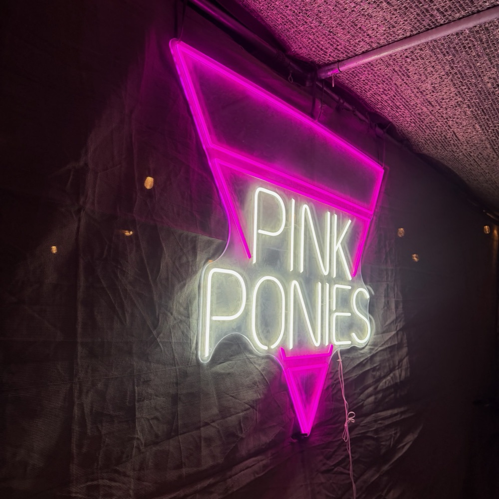 A neon sign at camp at night: "Pink Ponies" in white inside a glowing pink triangle pointing down, on a dark tent wall
